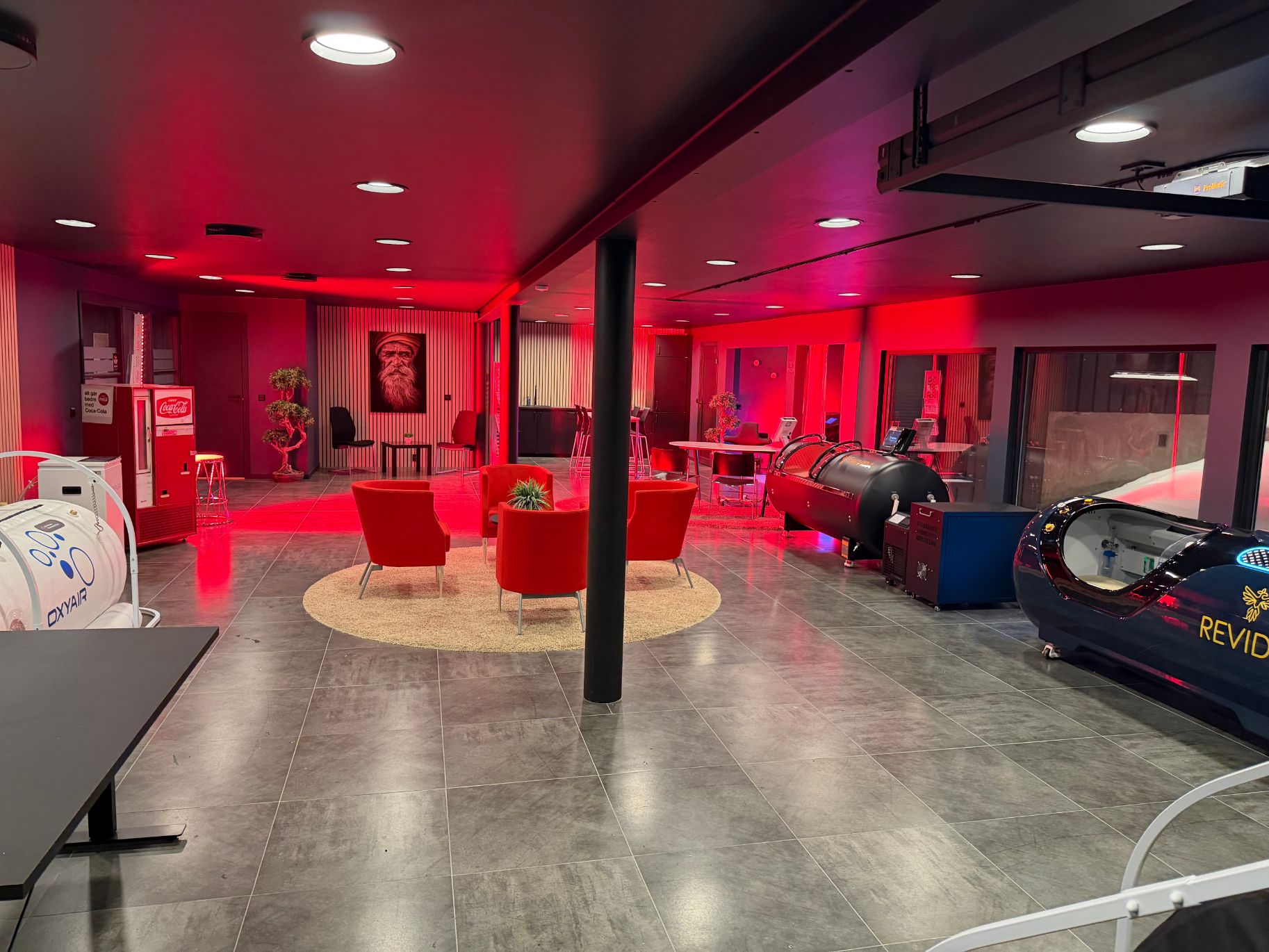 Åpent behandlingslokale med røde lys, terapikapsler og loungeområde med røde stoler.